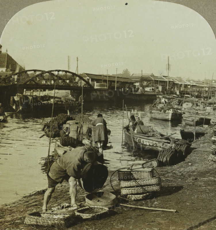 People and boats along a river in Yokohama, A glimpse of the busy river life. Yokohama, Japan, stereograph, number: '55', number: '180.', copyright notice: 'Copyright 1902 by CH Graves.', photographer: T. Enami (possibly), Yokohama, publisher: Universal Photo Art Company, Philadelphia, 1902, North America, Kanagawa, Honshu, paper, pasteboard, baryta paper, gelatin silver print, developing gelatin silver print, height 89 mm x width 178 mm, river, A riverside scene showing a group of people working near small wooden boats tied along the water, with a low arched metal bridge spanning the river in the background and a row of low buildings and poles along the far bank; in the foreground a person is bent over lifting a round container while other workers stand and sit on boats and a narrow dock handling baskets and bundles, with woven baskets and circular coiled mats stacked on the muddy shore and various poles and ropes scattered around, the water reflecting parts of the boats and bridge.