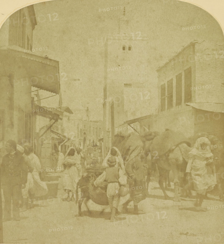 Street scene in an Arab city, possibly in Morocco, stereograph, inscription: 'C', number: '25175', inscription: 'SPECIAL ADDRESS FOR GOOD STEREOSCOPES, AND BEAUTIFUL STEREOSCOPIC PHOTOS.', photographer: August Frederik Willem Vogt, Morocco (possibly), 1897 - 1922, North Africa, pasteboard, paper, albumen print, height 88 mm x width 179 mm, street, A faded sepia street scene showing a narrow urban thoroughfare flanked by multi-story buildings with shuttered windows and awnings, a tall tower rising in the background, and many pedestrians wearing loose draped garments and head coverings moving along the street; some figures lead or ride animals, while others carry bundles or push carts, and market stalls with angled canopies extend from the building fronts.