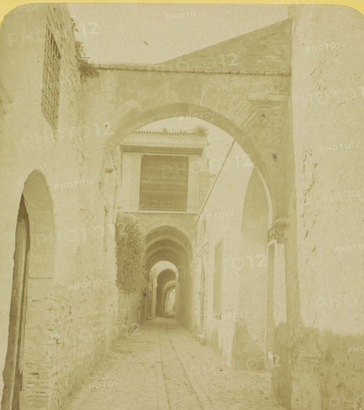 City gate in Tunis, Tunis. Porte arabe. Tunisie, stereograph, number: '28523', number: '46', photographer: JG Beers, Tunis, publisher: Nederlandsche Stereoscoop Maatschappij, Amsterdam, 1873 - 1890, North Africa, pasteboard, paper, albumen print, albumendruk, height 87 mm x width 179 mm, city gate, A narrow, pale-toned corridor framed by a series of stone arches receding into the distance, with textured walls on either side showing patches of rough masonry and worn plaster, a tiled pathway beneath, small recessed openings and windows along the walls, a rectangular barred opening visible above one arch, climbing vegetation clinging to parts of the left wall, and soft, diffused lighting creating long shadows and muted contrasts.