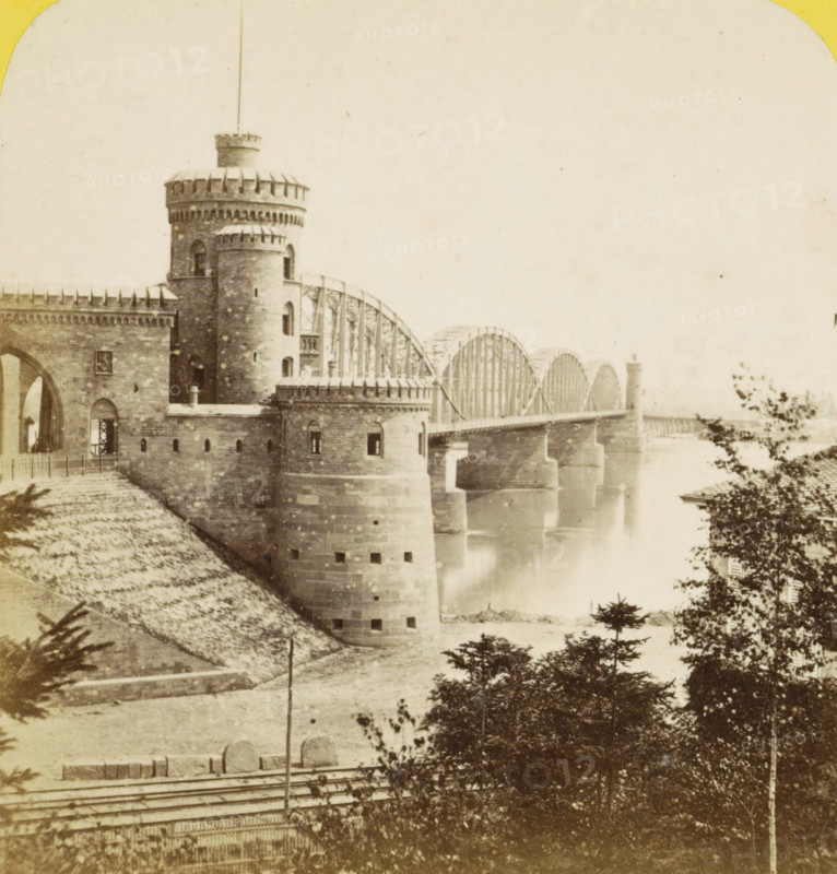 View of the Kaiserbrücke, now the Nordbrücke, in Mainz, Germany, Mayence (Hesse-Darmstadt), vue generale du pont fixe, prize de la nouvelle promenade, Allemagne, stereograph, inscription: 'J 60 cts', number: 'N. 423', inscription: 'Déposé', photographer: Hippolyte Jouvin, Mainz, 1860 - 1865, Rhineland-Palatinate, pasteboard, paper, albumen print, height 86 mm x width 176 mm, bridge, Kaiserbrücke, A sepia-toned photograph shows a stone fortress-like structure with rounded towers and crenellated parapets extending along a riverbank, connected to an arched metal bridge with multiple large semicircular trusses spanning the water; the river beneath the bridge appears calm and reflective, and the foreground includes sloped embankments, trees, and foliage partially obscuring the lower right area of the scene.