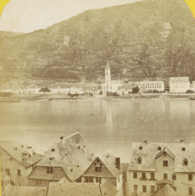 Saint-Goarshausen et le Rhin, vus de Saint-Goar, Sankt Goar and the Rhine, seen from Sankt Goarshausen, Germany, Allemagne, Bords du Rhin, stereograph, inscription: 'Déposé.', number: 'N. 483', photographer: Hippolyte Jouvin, Sankt Goarshausen, 1860 - 1870, Rhineland-Palatinate, pasteboard, paper, albumen print, height 86 mm x width 177 mm, river, city-view, and landscape with man-made constructions, Sankt Goar, A faded sepia photograph showing a riverside town with rows of buildings and steep tiled roofs in the foreground, a wide calm river in the middle reflecting light, and a distant shoreline with a cluster of larger buildings including a church with a tall steeple; behind them a steep terraced hillside rises, its textured slopes and horizontal lines visible under a pale sky.