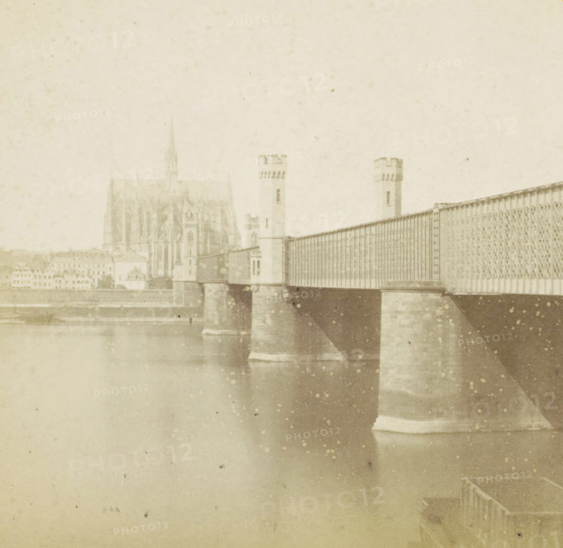 Pont fixe et Cathédrale, Dombrücke over the Rhine and Cologne Cathedral, Germany, Cologne (Prusse), Allemagne, stereograph, inscription: 'Déposé.', number: 'N. 543', inscription: '(Déposé)', photographer: Hippolyte Jouvin, 1864, North Rhine-Westphalia, pasteboard, paper, albumen print, height 86 mm x width 177 mm, church (exterior), bridge, Cologne Cathedral, Dombrücke, A faded sepia photograph shows a large iron bridge spanning a river, supported by stone piers rising from the water, with patterned metal railings and towers with crenellated tops at the near end; behind the bridge a Gothic cathedral with a tall pointed spire and detailed façade is visible above a row of buildings along the riverbank, and the water in the foreground reflects the bridge piers and has scattered spots and discolorations across the image.