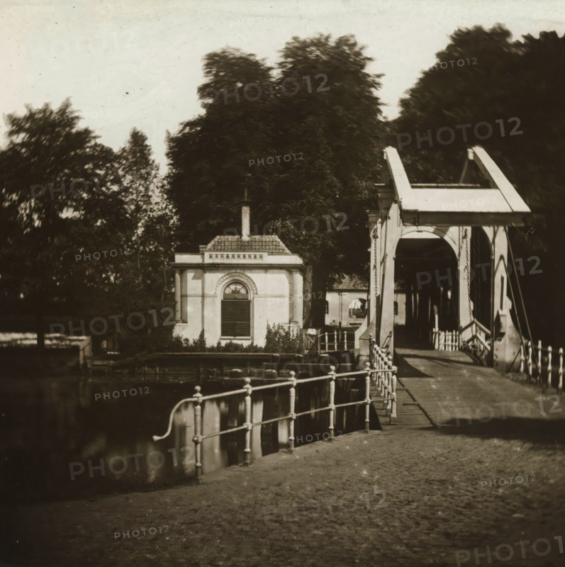 View of 's Gravelandsche poort in Weesp, stereoscopic photograph, address: 'NZ Voorburgwal, bij het Spui.', photographer: Pieter Oosterhuis, Weesp, 1856, Pieter Oosterhuis, photographer, inorganic material, glass, slide, height 85 mm x width 173 mm, bridge, A waterfront scene showing a small classical building with a tiled roof and a narrow chimney, an arched window with shutters on its facade, dense dark trees rising behind it, and a white wooden drawbridge with curved support beams and hanging chains spanning a canal; in the foreground a metal railing runs along the canal edge above water, a paved walkway leads toward the bridge, and reflections appear in the water beside the quay.