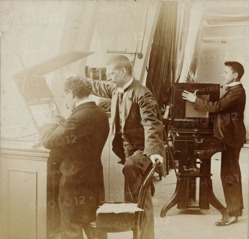 Photographers at work in a studio, stereograph, number: '12', Netherlands, 1882 - 1920, photographer, paper, pasteboard, daylight gelatin silver print, height 88 mm x width 179 mm, photographer at work, A sepia photograph showing three men in a room with tall windows and heavy curtains; one man seated at a window holding a magnifying glass to a flat surface, another man standing beside him leaning forward with one hand resting on the seated man's shoulder and the other on the back of a chair, and a younger man standing near a large wooden camera or box apparatus with his hands on its side. The men wear dark suits with high collars and cravats, and the room contains wooden furniture including a turned chair and a stand supporting the camera, as well as cylindrical containers on a shelf and visible window frames and moulding. The lighting is bright from the left, casting soft highlights and shadows across the scene, and the image has an aged, scratched texture.