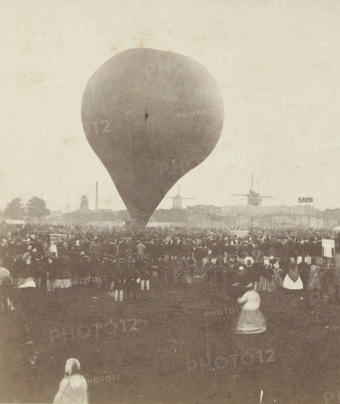 Ascent of Nadar's hot air balloon 'le Géant' on September 14, 1865, Giant balloon 'le Géant' by Nadar is launched in Amsterdam, September 14, stereograph, inscription: 'The Govert Flinckstraat has been cleared on this site., Amsterdam.', retailer's mark: 'BOOKSTORE, JAN D. BROUWER., KALVERSTRAAT E No 37, AMSTERDAM.', number: '10', photographer: Pieter Oosterhuis, Amsterdam, publisher: Jan Dirk Brouwer, 1865-09-14, pasteboard, paper, albumen print, height 85 mm x width 176 mm, balloon (aviation), A large teardrop-shaped balloon fills the center of the scene, its surface smooth and slightly mottled; a dense crowd of people stands and sits across the middle ground, with some figures closer to the foreground visible from the back and wearing long skirts or coats; in the distant background there are silhouettes of windmills and a few tall structures, and a long low fence or row of tents stretches horizontally; the overall image has a sepia tone and an aged photographic texture with faint sp...