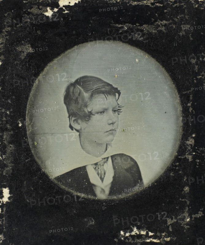 Portrait of an unknown boy, photograph, 1840 - 1860, photographer, metal, inorganic material, copper (metal), glass, paper, daguerreotype, in paper passe-partout, height 25 mm x width 24 mm, height 39 mm x width 33 mm x thickness 1.5 mm, historical persons, boy (child between toddler and youth) (sideview, profile), A faded oval photograph shows the head and upper torso of a young person with short, wavy hair and a serious expression, dressed in a dark jacket over a light shirt with a patterned necktie, surrounded by a dark, textured mat with scuffs and spots.
