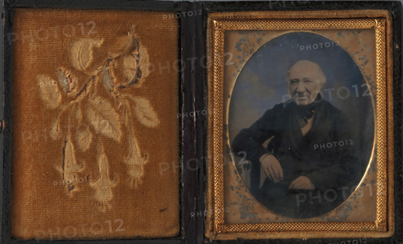 Portrait of an unknown man, photograph, c. 1855 - c. 1870, photographer, plant material, textile materials, biological material, inorganic material, glass, metal, velvet (fabric weave), wood (plant material), leather, ambrotype, in wooden case covered with leather with velvet lining, height 55 mm x width 44 mm, height 74 mm x width 62 mm x thickness 15 mm x width 126 mm, historical persons, sitting figure, A rectangular case opened to show two panels, the left panel lined with worn golden-brown velvet embossed with a faded floral design, the right panel containing an oval photograph encased in a decorative gold-colored frame with textured border and small ornamental corners; the photograph shows an older man seated, wearing a dark jacket and scarf, with one arm resting on an armrest and the other hand visible near his lap, his hair light and receding and his expression neutral, while age spots and speckles are visible on the photographic surface and the surrounding mat.
