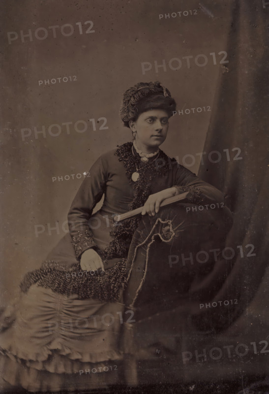 Portrait of an unknown woman with a hat, photograph, 1855 - 1900, photographer, metal, inorganic material, iron (metal), ferrotyping, height 104 mm x width 69 mm, historical persons - BB - woman, head-gear: cap, A seated woman wearing a dark dress with textured trim and a high collar is shown against a plain backdrop, she has an ornate hat with layered fabric and a decorative element, her hair is styled beneath it and she wears earrings and a brooch at her throat, one arm rests on an upholstered armrest with visible seam details while the other hand holds a slender folded object, the lower part of her dress shows gathered and layered fabric with ruffled edges, the image has a subdued monochrome tone and scattered surface blemishes.