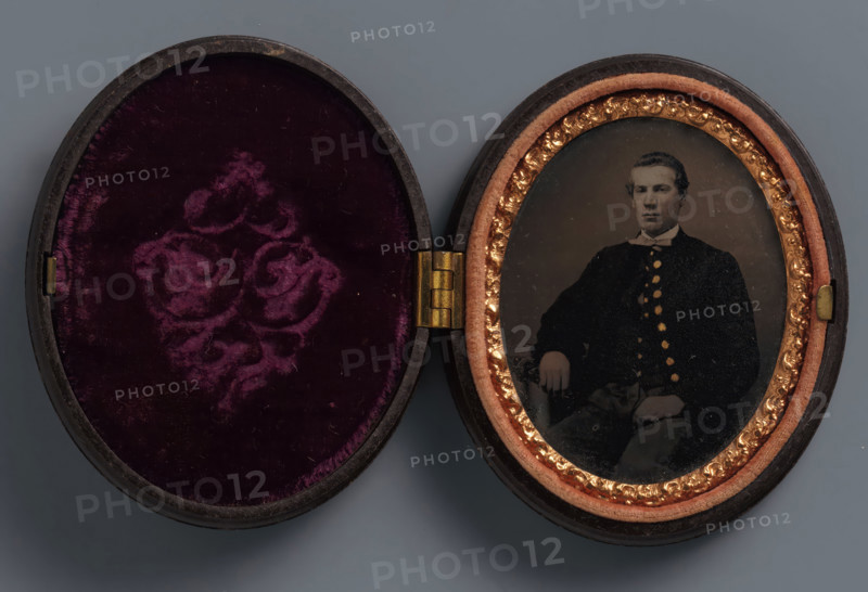 Portrait of an unknown man, possibly a soldier, photograph, union case, 1861, photographer, textile materials, organic material, inorganic material, metal, glass, velvet (fabric weave), plastic, ferrotyping, height 68 mm x width 56 mm x thickness 17 mm, anonymous historical person depicted, An open oval case with a hinged center showing a mirrored pair of interiors, the left side lined with deep purple velvet featuring a raised floral pattern and a dark rimmed edge, and the right side containing an oval photographic plate framed by a peach-colored padded border and a decorative gold inner trim, the photograph showing a seated man with short hair wearing a dark buttoned coat and a light-colored bow at the collar, his hand resting on his knee and the image surface showing small specks and slight wear.