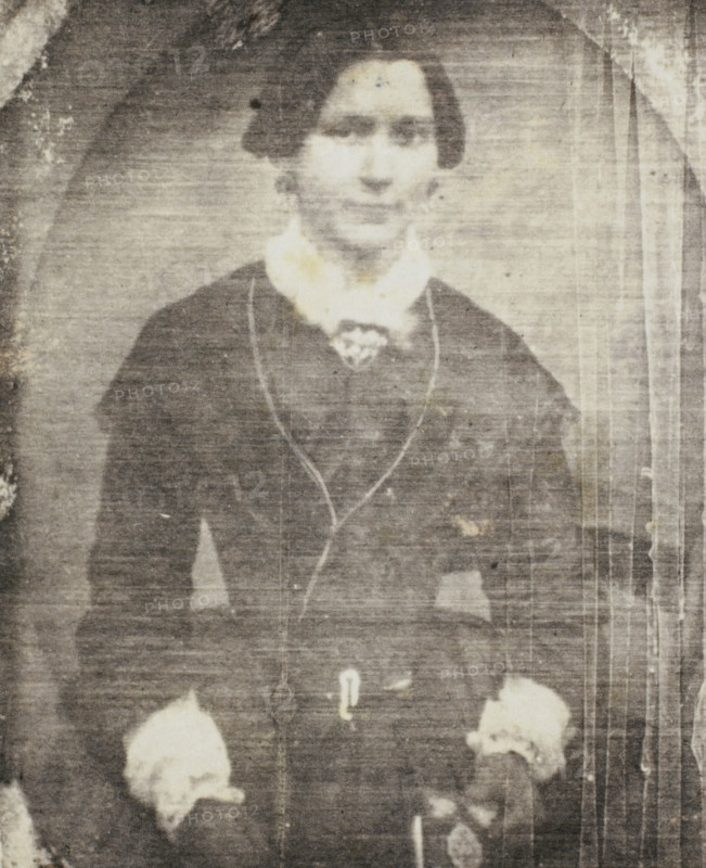 Photo reproduction of a daguerreotype of a portrait of an unknown woman, photograph, reproduction, France, after photo by anonymous, 1840 - 1860, photographer, paper, salted paper print, height 72 mm x width 59 mm, adult woman, historical persons not known by name - BB - woman, A faded black-and-white portrait of a person from the waist up wearing a dark dress or coat with a light-colored high collar and decorative trim, with hands resting at the sides holding light-colored floral objects, a faint oval backdrop around the head, and vertical streaks and spots from age or wear across the image.