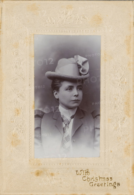 Christmas greeting with portrait of an unknown woman with a hat, photograph, Christmas card, Portrait of an unknown woman with a hat with the text 'With Christmas Greetings'., inscription: 'With, Christmas, Greetings', England, 1890 - 1910, photographer, paper, pasteboard, baryta paper, gelatin silver print, pressing, developing gelatin silver print, height 95 mm x width 67 mm, height 60 mm x width 33 mm, adult woman, anonymous historical person depicted, A sepia-toned portrait of a woman set within a textured ornate frame, showing her from the chest up wearing a tailored jacket with puffed sleeves, a high-collared blouse, and a patterned necktie; she wears a hat with a rounded crown and a decorative folded ribbon on one side, and her hair is styled back from her face; her expression is neutral as she looks slightly away from the camera; the frame around the photograph has embossed floral motifs and wavy textured patterns and shows some areas of discoloration.