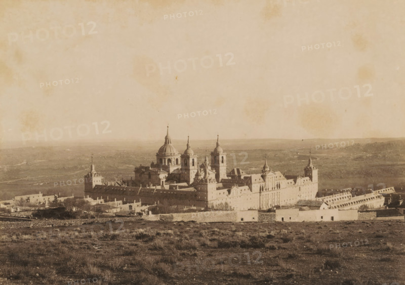Escorial, el Monasterio desde la presa del Romeral, View of the Escorial near San Lorenzo de El Escorial, photograph, number: '2.', photographer: C. Huerta, San Lorenzo de El Escorial, 1860 - 1900, Comunidad de Madrid, pasteboard, paper, albumen print, height 316 mm x width 367 mm, abbey, monastery, convent, Roman Catholic church, palace, mausoleum, A wide, sepia-toned panorama showing a large stone building complex with multiple towers, domes, and spires centered in the middle ground, surrounded by low walls and smaller structures; the foreground consists of dry, grassy terrain with scattered shrubs and uneven ground; the distant background shows a flat, open landscape with faint hills along the horizon; the sky above is pale with mottled stains and spots.