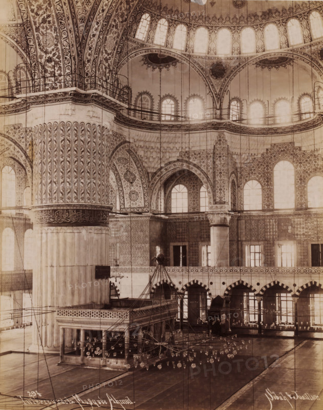 Intérieur de la Mosquée Ahmed, Interior of the Sultan Ahmet Mosque in Istanbul, photograph, number: '294', photographer: Sébah & Joaillier, Istanbul, 1888 - 1900, paper, albumen print, albumendruk, height 210 mm x width 264 mm, temple, shrine, Islam, Mohammedanism, Sultan Ahmet Mosque, The image shows an ornate interior space with large decorative columns and arches, intricate floral and geometric patterns covering walls and vaulted ceilings, rows of tall arched windows letting in light, suspended chains and many hanging glass lamps forming a chandelier arrangement, a raised ornate wooden or stone platform with carved balustrades, tiled lower walls with repeating motifs, a series of archways along a gallery, and polished floor surfaces reflecting light.