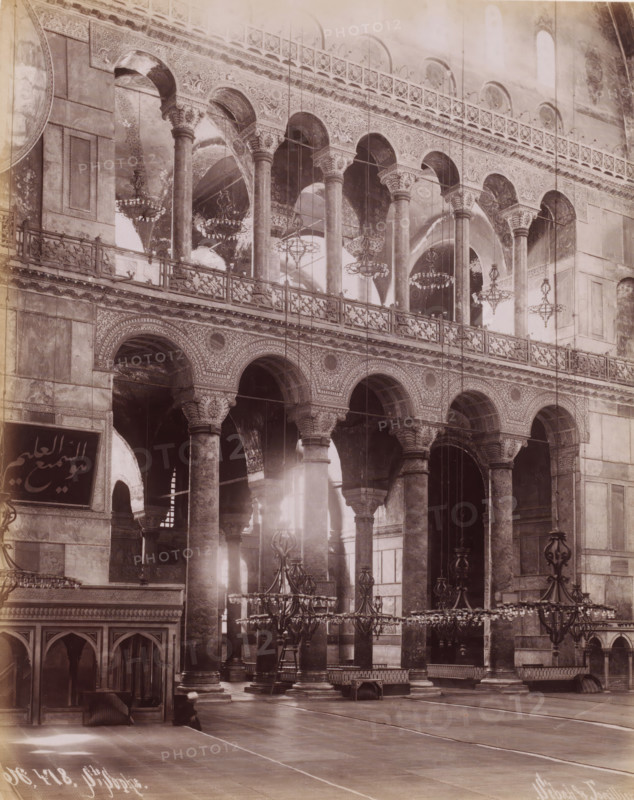 Interior of the Hagia Sophia in Istanbul, Ste Sophie, photograph, number: 'N. 478.', photographer: Sébah & Joaillier, Istanbul, 1888 - 1900, paper, albumen print, height 269 mm x width 210 mm, temple, shrine, Islam, Mohammedanism, Hagia Sophia, A large interior space framed by rows of tall stone columns with ornate capitals supporting a series of arched openings and an upper gallery with decorative balustrades, intricate carved patterns along the arches and spandrels, multiple hanging chandeliers of varying designs suspended at different heights, high vaulted ceilings with visible archways and painted or textured surfaces, a polished floor with visible seams, low wooden partitions and archways at one side, a few benches and raised platforms scattered near the columns, and soft light filtering through the openings creating highlights and shadows across the architectural details.