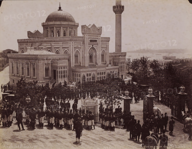 Ceremony of Islam, Crowd during the reception of Sultan Abdülhamit II for the Salat al-djuma (Friday afternoon prayer) at the Yildiz Hamidiye Mosque in Istanbul, photograph, number: '689', photographer: Abdullah Frères, Istanbul, 1876 - 1909, paper, albumen print, height 200 mm x width 259 mm, temple, shrine, Islam, Mohammedanism, crowd, mob, Yildiz Hamidiye Mosque, A large ornate building with a domed roof and a tall slender tower stands on a raised terrace overlooking a body of water, its façades featuring arched windows, decorative cornices, and latticework panels. In front of the building a crowd of uniformed men is gathered along a gated entrance and the terrace steps, some standing in formation while others cluster near carriages and horses. Trees and shrubs frame parts of the scene, and lamp posts with spherical glass tops appear near the entrance. Boats and distant shoreline structures are visible across the water behind the building, and the overall image has a sepia tone.