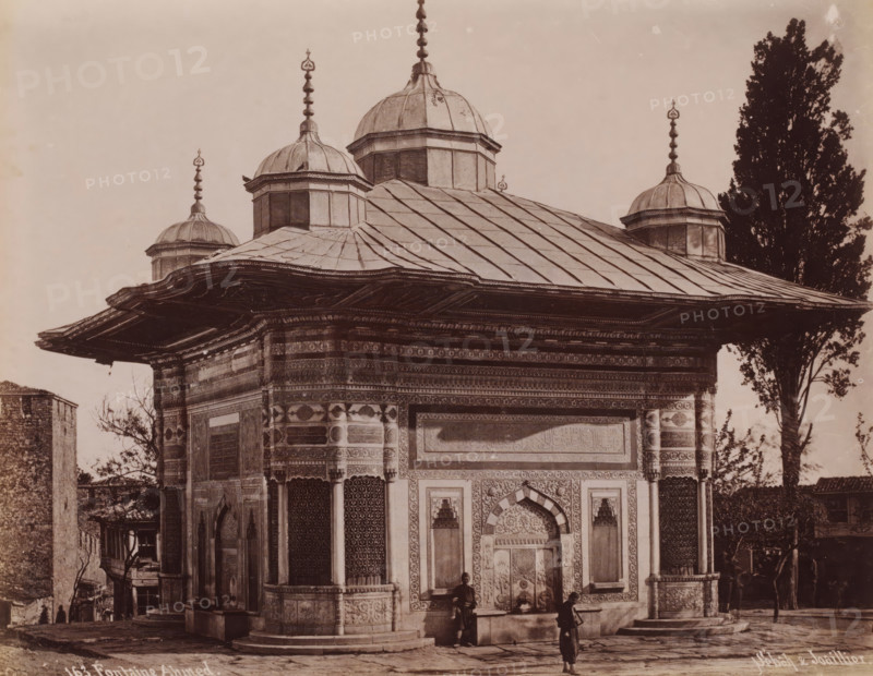 Fountain of Sultan Ahmed III in Istanbul, Fontaine Ahmed, photograph, number: '163.', photographer: Sébah & Joaillier, Istanbul, 1888 - 1900, paper, albumen print, albumendruk, height 207 mm x width 267 mm, utilitarian, public pump or water fountain, Topkapi Palace, Fountain of Sultan Ahmed III, A stone building with ornate carved panels and patterned tile work has a wide overhanging roof and multiple small domes topped with finials; the roof shows segmented metal panels and decorative edges, and the facade features arched doorways, recessed windows with lattice screens, and detailed relief borders; two figures stand near the entrance, one closer to the doorway and another a short distance away holding a stick or pole, and a tall tree rises beside the building while another smaller tree and adjacent structures are visible at the sides.