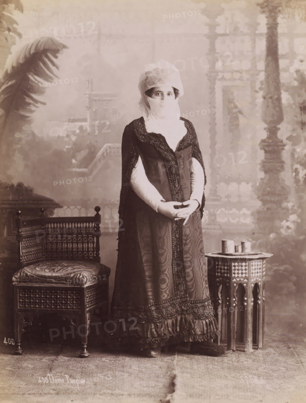 Portrait of an unknown Turkish woman, Dame Turque, photograph, number: '400', photographer: Jean Pascal Sébah, Middle East, 1888 - 1900, paper, albumen print, height 270 mm x width 210 mm, adult woman, anonymous historical person portrayed, A sepia-toned portrait of a person standing between an ornate wooden chair and a small decorative table, wearing a long dark dress with patterned trim and a light-colored head covering that also veils the lower face, with visible eyes looking toward the camera; the person’s hands are clasped at waist level over the dress, and the outfit includes long light-colored sleeves beneath the dark outer garment; the chair has carved latticework and an upholstered cushion with a decorative pattern, while the small table has a geometric inlaid design and holds three cylindrical containers; the backdrop shows a painted scene with architectural and foliage motifs and a large palm frond visible at the left edge of the image.