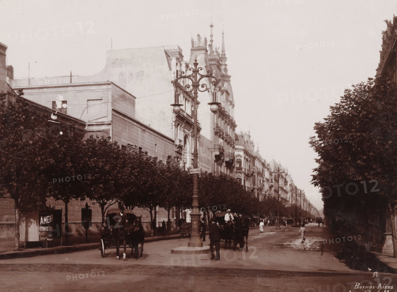 Buenos-Aires, Avenida de Mayo, View of Avenida de Mayo in Buenos Aires, photograph, monogram: 'AC', inscription: 'Buenos Aires 1904, 5', photographer: anonymous, Buenos Aires, 1904 - 1905, paper, pasteboard, baryta paper, daylight collodion silver print, height 166 mm x width 225 mm, height 259 mm x width 334 mm, city-view, and landscape with man-made constructions, A wide city street lined with tall, ornate buildings on one side and rows of trimmed trees on both sides; a decorative lamppost with curved metalwork stands near the center foreground, flanked by horse-drawn carriages and people walking or standing on the wet road; the facades of multi-story buildings show balconies, sculpted details, and steep rooflines with spires, while the tree canopies form a dark, leafy border along the avenue and cast shadows onto the sidewalk.