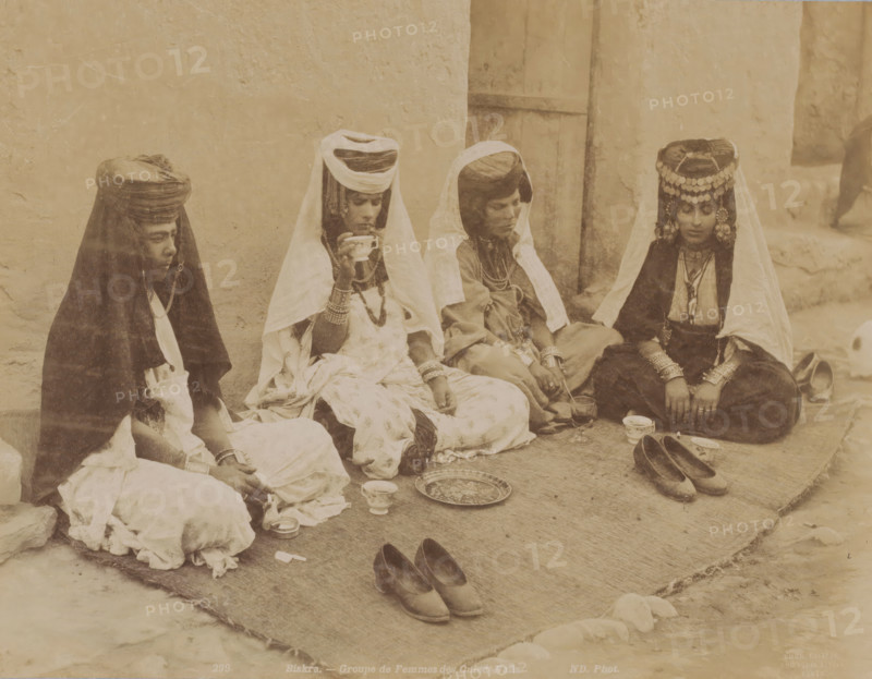 Biskra, groupe de femmes des Ouled-Naïls, Group portrait of four unknown Ouled Naïl women in Algeria, photograph, number: '299', photographer: Neurdein Frères, Algeria, 1863 - 1900, North Africa, pasteboard, paper, albumen print, height 278 mm x width 345 mm, anonymous historical persons depicted in a group, in a group-portrait - BB - woman, Four seated women wear layered garments and head coverings, with two wearing veils that drape over their shoulders and one wearing a decorative headpiece with hanging ornaments; they sit on a woven mat against a plastered wall and a wooden door, with various cups and a patterned tray placed on the mat in front of them and pairs of shoes positioned nearby.