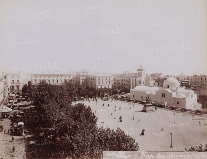 View of the place du Gouvernement (now the place des Martyrs) with the Djama?a el Djedid in Algiers on the right, Alger, place du gouvernement, photograph, number: '2330 ?', Algiers, 1860 - 1900, photographer, North Africa, paper, albumen print, height 188 mm x width 242 mm, temple, shrine, Islam, Mohammedanism, square, circus, Djama?a el Djedid, A wide sunlit plaza bordered by multi-story buildings and a row of trees sits in the lower part of the image, with people scattered across the open paved area and a few horse-drawn carriages and carts along the edges; a domed white building with smooth curved roofs and small arched windows occupies the right foreground, adjacent to a tower featuring a square face and circular decorative elements, while a statue on a pedestal stands near the dome and a street lined with shaded trees and parked carriages runs along the left side of the scene.