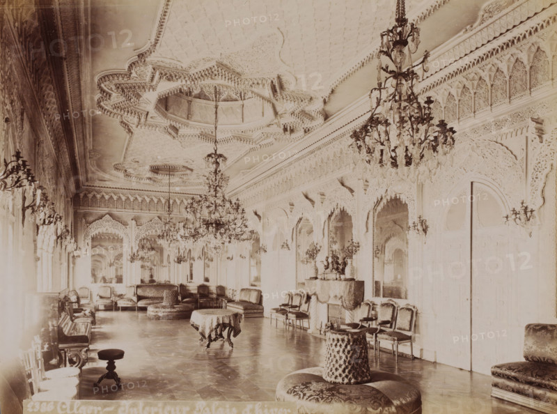 Alger, interior Palais d'hiver, Interior of the Dar Hassan Pacha in the Kasbah of Algiers, photograph, number: '2386', Kasbah of Algiers, 1860 - 1900, photographer, North Africa, Algiers, paper, albumen print, height 189 mm x width 250 mm, palace, Dar Hassan Pacha, A long ornate room with a high carved ceiling and multiple decorative chandeliers suspended along its length, walls covered with intricate carved arches and ornamental moldings, rows of upholstered chairs and sofas arranged along the sides, several round cushioned ottomans and small tables draped with cloths placed on a polished floor, wall sconces with hanging ornaments, a console table with decorative vases and objects beneath a mirror, and a large carved door set into one wall.