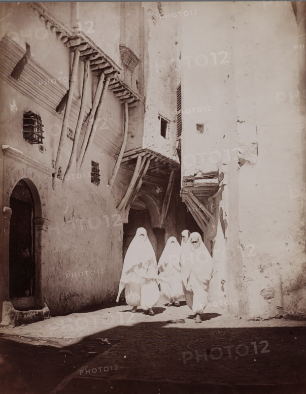 Alger (Rue Arabe), Four unknown veiled women on the street in Algiers, photograph, number: '2018.', Algiers, 1860 - 1900, photographer, North Africa, paper, albumen print, height 244 mm x width 187 mm, adult woman, street, A narrow sunlit alley flanked by tall, weathered buildings with rough plaster walls and projecting wooden beams, showing small barred windows and an arched doorway on the left, and timbered overhangs and recessed openings above; in the foreground a group of figures draped in light-colored garments covering their heads and bodies walk toward the camera, their faces mostly obscured, casting shadows on the cobbled ground.