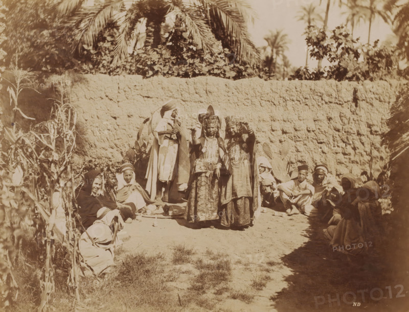 Danse des Ouled Naïls, Group Ouled Naïl people in Algeria, Types Algeriens, photograph, number: 'L. 259', photographer: Neurdein Frères, Algeria, c. 1863 - 1900, North Africa, pasteboard, paper, albumen print, height 278 mm x width 345 mm, anonymous historical persons depicted in a group, in a group-portrait, A group of people arranged outdoors in front of a textured earthen wall with tall palm fronds and foliage visible above it, with two women standing near the center wearing ornate headdresses and long layered garments while other figures sit or crouch around them on the ground, some wrapped in cloaks or head coverings, with a person standing slightly behind the central pair wearing a draped robe and headwrap and other seated individuals forming a loose semicircle, dried plants and stalks along the left edge, scattered pottery or small vessels on the ground, and patches of sunlit bare earth and grassy tufts in the foreground.