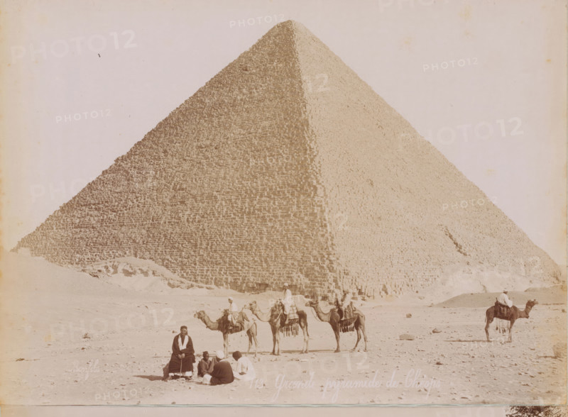 View of the Pyramids of Giza, photograph, Giza, 1860 - 1900, photographer, North Africa, pasteboard, paper, albumen print, height 190 mm x width 278 mm, height 396 mm x width 290 mm, pyramid (historical grave form), Pyramids of Giza, A large stone pyramid fills the background with its stepped blocks visible on two faces, and a sandy foreground shows a group of people and camels; some riders sit atop camels wearing loose garments and head coverings while others sit on the ground in a small cluster, and one person sits alone facing the camera.