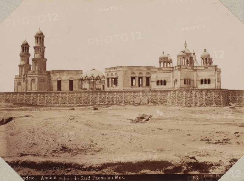 Alexandrie, ancien palais de Saïd Pacha au Mex, View of the palace of Mohammed Said at the port of Alexandria, photograph, number: '93.', photographer: Luigi Fiorillo, Alexandria, 1870 - 1890, North Africa, pasteboard, paper, albumen print, height 316 mm x width 404 mm, Person, Mohammed Said (governor of Egypt), palace, Palace of Mohammed Said, A wide, sepia-toned photograph shows a large stone building complex behind a textured stone wall, with multiple domes and ornate towers rising above the roofline; two tall cylindrical towers with open-arched upper sections stand on the left, while a variety of rounded domes and small cupola-like structures appear toward the right, some supported by slender columns. The façade of the buildings features rectangular and arched openings, some appearing dark and empty, and decorative circular and arched window frames on portions of the walls. In front of the wall is a barren, uneven expanse of ground with scattered low vegetation and shallow depressi...