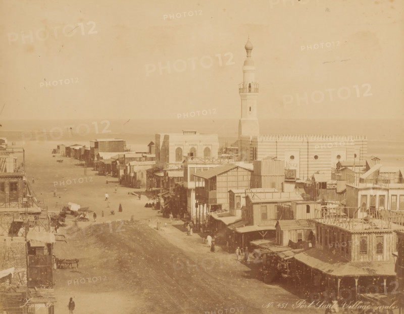 Port Saïd Village arabe, View of Port Said, photograph, inscription: 'photo Zangaki, Port Said Arab village', number: 'No. 451', photographer: Zangaki, C. & G., Port Said, 1870 - 1910, North Africa, pasteboard, paper, albumen print, height 220 mm x width 278 mm, height 348 mm x width 454 mm, public road in village, temple, shrine, Islam, Mohammedanism, A sepia-toned street scene with rows of wooden and masonry buildings lining both sides of a wide unpaved road that leads toward a shoreline, a tall minaret rising above a large striped building near the water, small awnings and balconies projecting from many structures, scattered groups of people walking or standing along the street, carts and market stalls visible near the foreground, and the calm sea forming a horizontal band on the horizon.