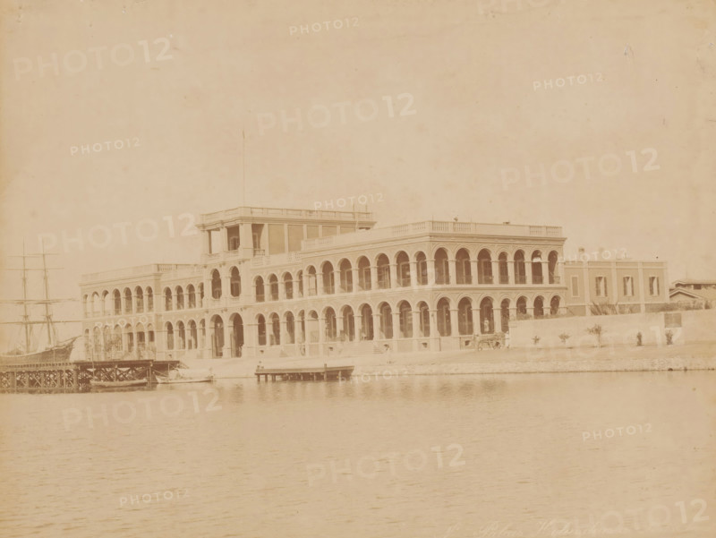Port office, called the Palais Hollandais, in Port Said, Palais Holandais à Port Saïd, photograph, inscription: 'Port Said', photographer: Zangaki, C. & G., Port Said, 1870 - 1910, North Africa, pasteboard, paper, albumen print, height 212 mm x width 276 mm, height 346 mm x width 453 mm, harbour, A sepia-toned photograph showing a large waterfront building with two levels of arched colonnades and a central raised section, a pier and small boats in front of the building, a multi-masted sailing ship visible on the left, a low wall and a row of small trees or shrubs along the shoreline to the right, and calm water occupying the foreground.