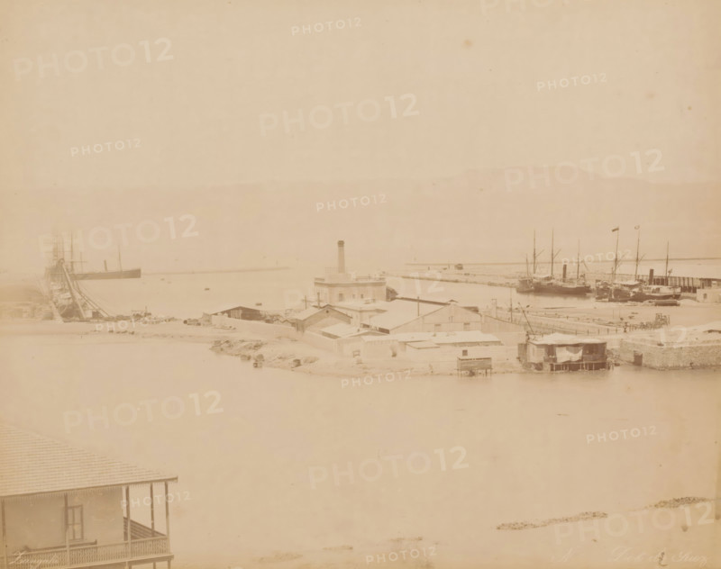 Dok de Suez, Dock at the Suez Canal, photograph, number: 'No', inscription: 'foto Zangaki, Suez', photographer: Zangaki, C. & G., Suez Canal, 1870 - 1910, North Africa, pasteboard, paper, albumen print, height 349 mm x width 454 mm, canal, dry-dock, floating-dock, A faded panoramic view of a harbor area showing a stretch of calm water bordered by low buildings and docks, a cluster of ships moored at the right with tall masts and flags, another large vessel at the left with visible rigging, a central blocky building with a tall chimney rising above surrounding single-story structures, assorted small sheds and waterfront platforms, and a foreground porch with a decorative railing partially visible at the lower left.