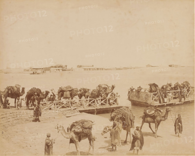 Ferry across the Suez Canal at Al-Qantarah, Egypt, Pont de la Syrie a Kantara, photograph, number: 'No', photographer: Zangaki, C. & G., Al-Qantarah Gharb, 1870 - 1910, North Africa, pasteboard, paper, albumen print, height 349 mm x width 454 mm, ferry, hoofed animals: camel, A wide sepia-toned waterside scene showing a sandy shore with a wooden pier extending into the water and a flat expanse of river or lake beyond, a low line of simple buildings and a few trees on the far bank, and numerous camels on the shore and on the pier loaded with large bundles and saddles; many men in traditional robes and head coverings stand or sit among the camels, some holding ropes or leading animals, while a few people are on the pier near a small boat or platform at the water's edge.