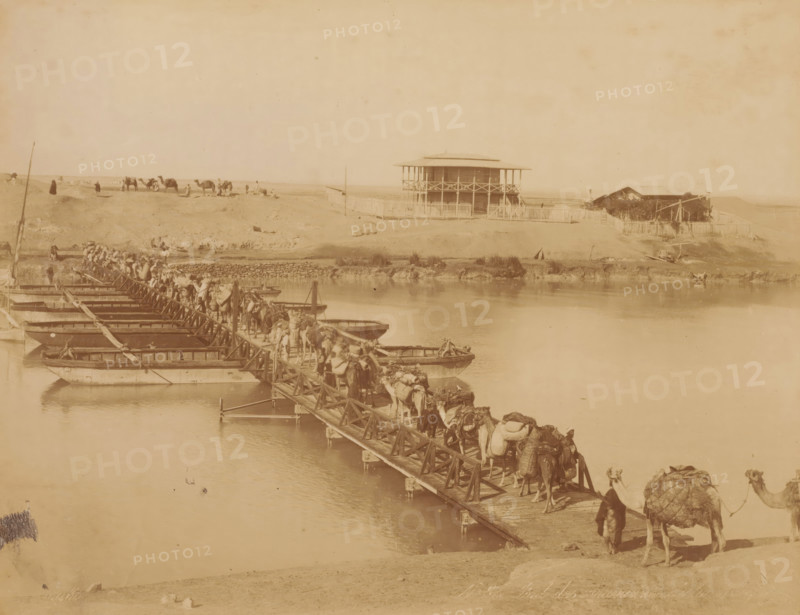 Caravan with camels or dromedaries on a pontoon bridge, Pont des caravannes de la ..., photograph, number: 'No ...', photographer: Zangaki, C. & G., 1870 - 1910, pasteboard, paper, albumen print, albumendruk, height 348 mm x width 454 mm, hoofed animals: camel, pontoon bridge, A sepia-toned riverside scene shows a long wooden pontoon bridge extending across calm water, with numerous camels and people walking along it; small flatboats are moored alongside the bridge, and cargo is visible on some camels. On the far bank a two-story wooden building with a wraparound balcony and a fenced yard stands atop a low rise, with a smaller structure and vegetation nearby. In the background a sandy landscape stretches with scattered figures and a group of camels standing on the plain. The sky appears clear and pale above the horizon.