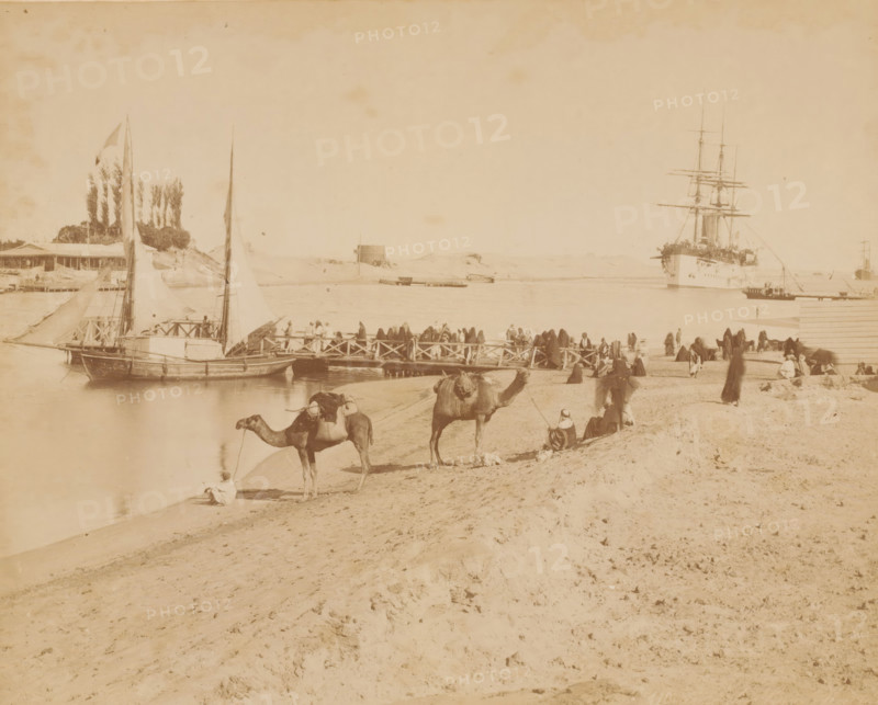 Pont de la Syrie à Kantara, Crossing point for caravans across the Suez Canal at Al-Qantarah, photograph, number: 'No. 110', photographer: attributed to Zangaki, C. & G., Al-Qantarah Gharb, 1870 - 1910, North Africa, Attributed to, pasteboard, paper, albumen print, albumendruk, height 219 mm x width 271 mm, height 348 mm x width 453 mm, canal, hoofed animals: camel, Suez Canal, A wide sandy shoreline with footprints and ridges extends across the foreground toward a busy riverbank where several camels stand or rest, some with saddles and packs, and nearby people sit or stand wearing flowing robes and head coverings; a wooden jetty projects into the water crowded with people, small sailboats moored alongside display tall triangular sails, and farther out a large multi-masted ship is anchored with rigging and spars visible against the sky, while low buildings, trees, and dunes line the opposite shore.