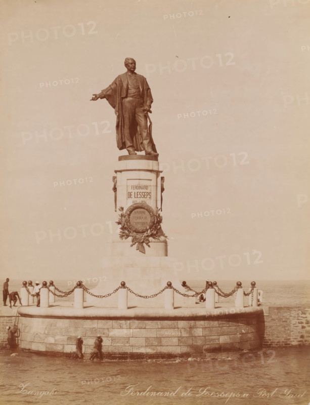 Statue of Ferdinand de Lesseps at the Suez Canal in Port Said, Ferdinand de Lesseps a Port Said, photograph, photographer: Zangaki, C. & G., Port Said, 1894 - 1910, North Africa, paper, albumen print, height 278 mm x width 215 mm, Person, Ferdinand de Lesseps, monument, statue, Suez Canal, A large bronze statue of a robed man stands atop a stone pedestal on a circular stone platform by the water, the figure shown with one arm extended and the other holding an object at his side; ornamental wreath or relief decoration is affixed to the front of the pedestal. The platform is surrounded by short stone bollards linked by heavy chains and a low curved stone wall, with a few people visible walking along the base of the platform near the water and a couple of figures near the bollards. The scene has a muted, sepia-toned appearance and an expanse of sky fills the upper portion of the image.