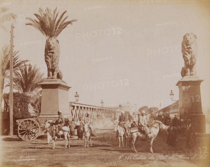 Entrance to the Qasr al-Nil Bridge, Qasr al-Nil Bridge over the Nile in Cairo, photograph, number: 'No 385', photographer: Zangaki, C. & G., Cairo (city), 1870 - 1910, Ottoman Empire, North Africa, paper, albumen print, height 223 mm x width 283 mm, bridge in city across river, canal, Qasr al-Nil Bridge, A wide sepia-toned scene shows a bridge flanked by two tall stone plinths each topped with a large lion statue, palm fronds rising behind the left lion and ornate street lamps mounted along the bridge. People and riders on donkeys and horses move across the bridge, some figures standing or walking, others seated on animals, and a wooden cart wheel is visible at the left foreground. Buildings and trees are visible in the distant background beyond the bridge, and the ground in the foreground appears dusty.