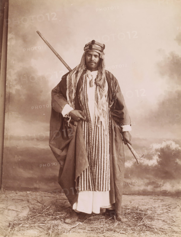 Costume Bedoin, Portrait of an unknown Bedouin man, photograph, number: 'N. 231', photographer: G. Lekegian & Co., Middle East, 1887 - 1900, paper, albumen print, height 212 mm x width 271 mm, anonymous historical person portrayed, wandering communities: nomads, A sepia-toned portrait of a man standing full-length against a painted backdrop showing clouds and distant landscape, wearing layered traditional garments including a long striped robe over a lighter ankle-length garment and a loose outer cloak, a patterned headscarf wrapped and secured with a band around his head, a tassel hanging at his chest, boots visible at his feet, a long staff resting diagonally over his shoulder, and a curved walking stick held in one hand, with straw scattered on the ground.