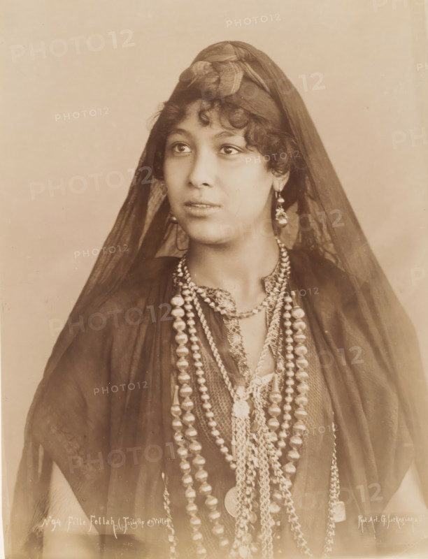 Fille Fellah (Toilette de Ville), Portrait of an unknown Fellah woman, photograph, number: 'No 94', photographer: G. Lekegian & Co., Middle East, 1887 - 1900, paper, albumen print, height 276 mm x width 212 mm, adult woman, anonymous historical person portrayed, A sepia-toned portrait of a young woman shown from the chest up wearing a dark sheer veil draped over her head and shoulders, a tied headscarf at the crown, curly hair visible at her forehead, large dangling earrings, multiple layered beaded necklaces of varying shapes and sizes, a patterned neckline beneath the necklaces, and a neutral plain background.