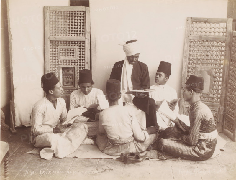Ecole Arabe de jeuns garçons, School class with pupils and teacher, photograph, number: 'No 91', photographer: G. Lekegian & Co., Middle East, 1887 - 1900, paper, albumen print, height 210 mm x width 275 mm, teacher and pupil (pupil), A group of young men sit closely together on a tiled floor in front of patterned wooden screens, each holding open books and wearing long tunics and tall cylindrical hats, while a central figure with a wrapped headscarf and darker outer garment sits slightly elevated and looks down at an open book in his hands; footwear lies on the floor nearby and the scene is framed by carved lattice panels against a plain wall.