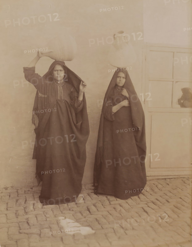 Double portrait of two unknown Egyptian women with jugs on their heads, Femmes Egypte Afalages ?, photograph, inscription: 'Dubbel', Middle East, 1860 - 1900, photographer, paper, albumen print, height 280 mm x width 219 mm, adult woman, Two women stand side by side against a plain wall and a panelled door, each wearing dark full-length garments with head coverings; the woman on the left holds a large vessel balanced on her head with one hand while lifting part of her head covering with the other, and the woman on the right also balances a vessel on her head while holding her garment closed with one hand; both wear necklaces and have neutral facial expressions, and cobblestones are visible beneath their feet.