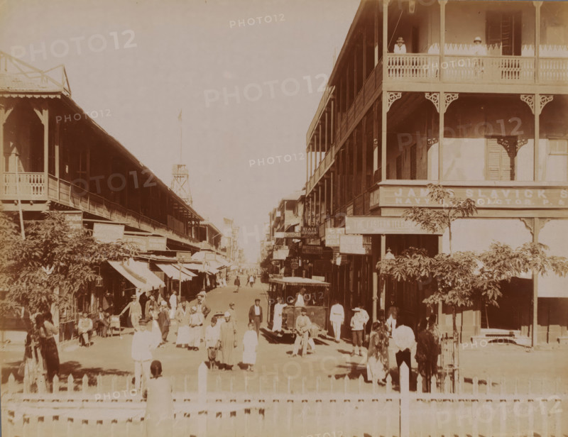 Rue du Commerce in Port Said, Rue du Commerce, Port Saïd, photograph, Port Said, 1860 - 1900, photographer, North Africa, paper, albumen print, height 215 mm x width 278 mm, street, A sepia-toned street scene shows two rows of tall wooden buildings with covered balconies and decorative railings flanking a wide avenue. People in period clothing walk and stand along the street and on the sidewalks, some grouped in small clusters and others moving toward the distance. A few horse-drawn carriages and a vehicle with an open cabin are visible among the pedestrians. Trees and awnings project from the buildings, casting shadows on the ground. Two figures stand on an upper balcony on the right building, and ornamental brackets accent the building corners. A low fence runs across the foreground.
