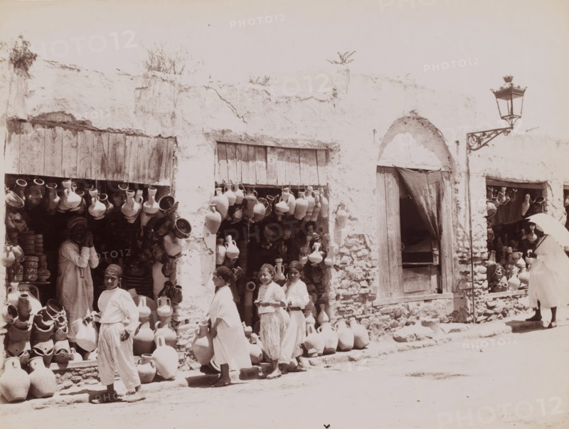View of a jar shop, photograph, inscription: 'illegible', Middle East, 1860 - 1900, photographer, paper, albumen print, height 199 mm x width 257 mm, shop, store, container of ceramics: jar, jug, pot, vase, A sunlit street scene showing a worn white plaster building with wooden shutters and an arched doorway draped by a dark curtain, walls marked by cracks and patches of exposed stone; numerous clay jugs and pottery of various shapes hang from rafters, sit on shelves, and are stacked along the sidewalk and in shop openings; several people in light, flowing garments stand and smile near the pottery displays, including a group of young figures in the foreground and an adult partially visible inside a shop; a tall ornate street lamp on a decorative bracket projects from the wall near the arched doorway; the ground in front of the shops is dusty.