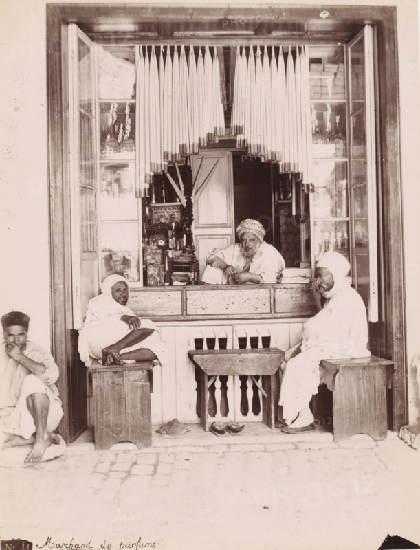 Perfume merchant, Perfume seller in his shop, presumably in Tunis, photograph, number: 'N. 44.', Tunis, 1860 - 1900, photographer, North Africa, paper, albumen print, height 259 mm x width 199 mm, shop, store, using scents, perfumes, ointments, A shopfront with an open window framed by wooden panels and a hanging vertical blind made of long thin rods, revealing an interior shelf filled with bottles and containers. A man leans on the counter inside, wearing a patterned headwrap and a loose garment, with his arms resting on the wooden ledge and a relaxed expression. Two other men sit on wooden stools outside the counter, both wrapped in light-colored draped robes and head coverings; one sits cross-legged with bare feet visible and an arm resting on his knee, the other sits facing slightly toward the counter with hands near his face. A small wooden table with a carved balustrade front stands between the stools, and a pair of sandals lie beneath it on the cobbled ground. The shop windows o...