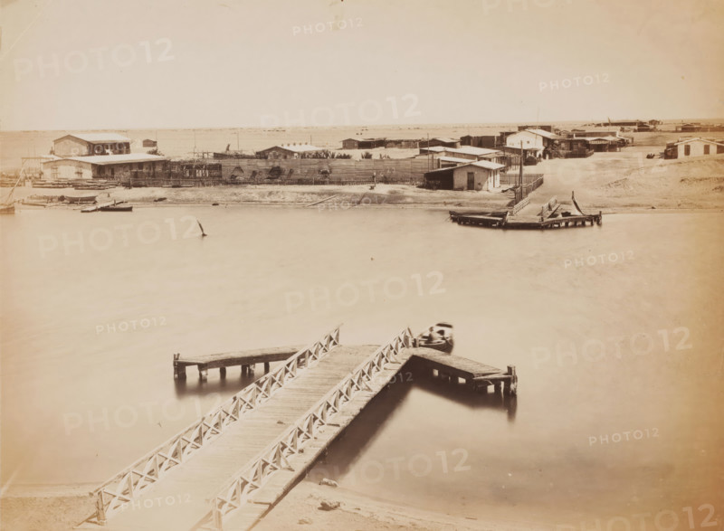 View of a ferry, possibly in Egypt, photograph, Egypt, 1860 - 1900, photographer, North Africa, paper, albumen print, height 202 mm x width 270 mm, ferry, A sepia-toned waterfront scene showing a wooden pier with crisscross railings extending from the foreground into the water and branching into smaller platforms, a small boat moored alongside one platform, a blurred boat near the pier, calm reflective water filling much of the frame, a sandy shoreline across the water lined with low wooden buildings and fencing, several simple houses and sheds clustered behind the fence, additional docks and small boats along the far shore, and an expansive pale sky above.