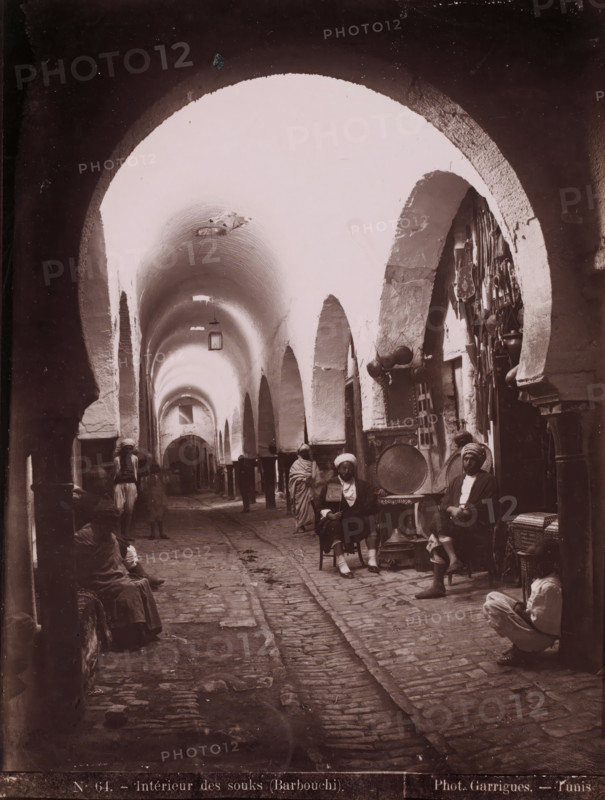 Intérieur des souks (Barbouchi), View of the Souk el-Trouk in the Medina of Tunis, on the right the shop of the Barbouchi Frères, photograph, number: 'N. 64.', photographer: J. André Garrigues, Medina of Tunis, 1860 - 1900, North Africa, Tunis, paper, albumen print, height 259 mm x width 198 mm, market, Souk el-Trouk, A covered stone passage with a vaulted ceiling and a series of arches along both sides, lined with small alcoves and shop openings, cobbled floor with a central narrow groove, hanging lanterns from the ceiling, several seated men wearing wrapped head coverings and robes positioned near the shop openings, metal trays and hanging objects displayed on the right side wall, a few standing figures further down the passage, and various low stools, baskets, and containers placed along the sides.