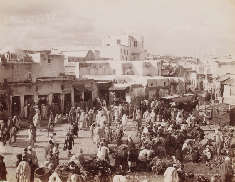Mds de Légumes, Greengrocers on a square, probably in Tunis, photograph, number: 'No 84', photographer: J. André Garrigues, Tunis, 1860 - 1900, North Africa, paper, albumen print, albumendruk, height 199 mm x width 255 mm, market, A crowded outdoor market scene in front of a cluster of low, light-colored buildings with flat roofs and small windows, with many people wearing long robes and head coverings milling about, standing, walking, and sitting among baskets, bundles of produce, and market stalls, while some vendors display goods on the ground and others work around carts and awnings, and a larger multi-story white building rises behind the row of shops, with a wide open sky above.