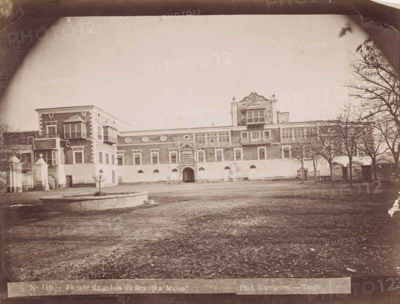 View of the Dar al-Taj in La Marsa, Façade du palais du Bey (la Marsa), photograph, number: 'N. 110.', photographer: J. André Garrigues, La Marsa, 1860 - 1900, North Africa, paper, albumen print, height 198 mm x width 258 mm, palace, Dar al-Taj, A wide sepia photograph of a large ornate building seen from an open courtyard, showing a long facade with decorative windows, balconies, and architectural details, with a central arched entrance and a raised ornate section above it, flanked by lower wings; a circular stone fountain basin sits in the foreground on bare ground, leafless trees line the right side near a wrought iron fence and gate, and a few small plants and architectural elements are visible near the left side of the building.