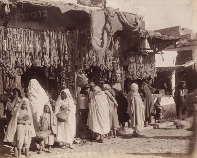 Grocery stores with customers, probably in Tunis, Epiciers Arabes, photograph, number: 'No 300', photographer: J. André Garrigues, Tunis, 1860 - 1900, North Africa, paper, albumen print, height 199 mm x width 248 mm, shop, store, A busy outdoor market scene showing a storefront with many ropes and bundles hanging from the awning and beams, fabrics draped over the top, and various goods displayed inside and below the hanging items; several people wearing long loose garments and head coverings stand and move in front of the shop, some carrying baskets or bundles, including adults and children; stone steps and a cobbled ground are visible beneath their feet; to the right a man wearing a cap stands near the building while a small child sits on a step holding an object; architectural elements of the shop and adjacent buildings, including rough walls and shaded recesses, appear in the background.