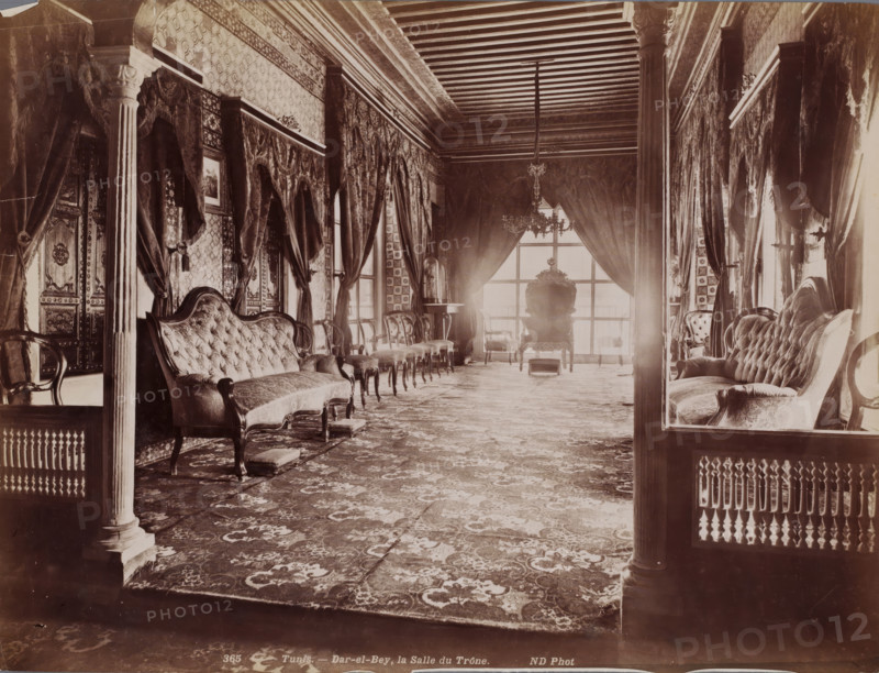 Tunis, Dar-el-Bey, la Salle du Trône, Throne Room of the Dar El Bey in the Medina of Tunis, photograph, number: '365', photographer: Neurdein Frères, Medina of Tunis, 1863 - 1900, North Africa, paper, albumen print, height 208 mm x width 273 mm, throne-room, Dar El Bey, A long, elaborately furnished room with patterned carpets and high ceilings featuring exposed beams, flanked by tall, ornate columns and heavy draped curtains along both sides; upholstered sofas and chairs with tufted backs line the walls, interspersed with small footstools and paired side chairs, while framed artwork and carved wooden panels adorn the walls; a large window at the far end is partially covered by heavy curtains and a central ornate armchair sits in front of it beneath a hanging chandelier, with additional small tables and decorative objects visible near the window.